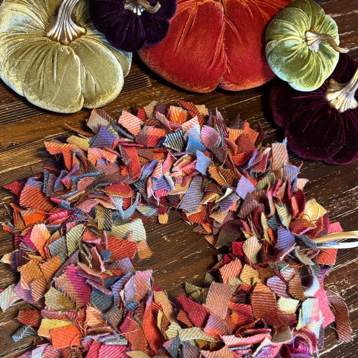 A handmade autumn-themed fabric fringe wreath made of multicolored plaid and striped fabric strips, displayed on a dark wood floor with decorative velvet pumpkins in the background.