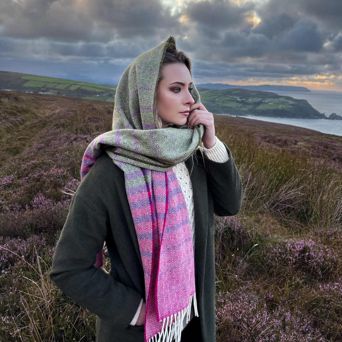 A woman wearing a green and pink stripe wool scarf and a dark green coat stands outdoors in hills of donegal on a grassy landscape with heather under a dramatic cloudy sky.