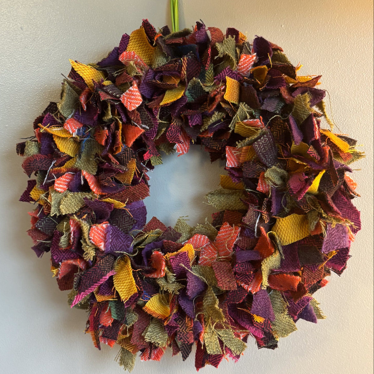 A handmade fabric slub yarn wreath hanging on a plain off-white wall. The wreath is made from many small pieces of textured fabric in autumnal colors including purple, orange, yellow, and brown, with a small lime green hanging tag at the top.