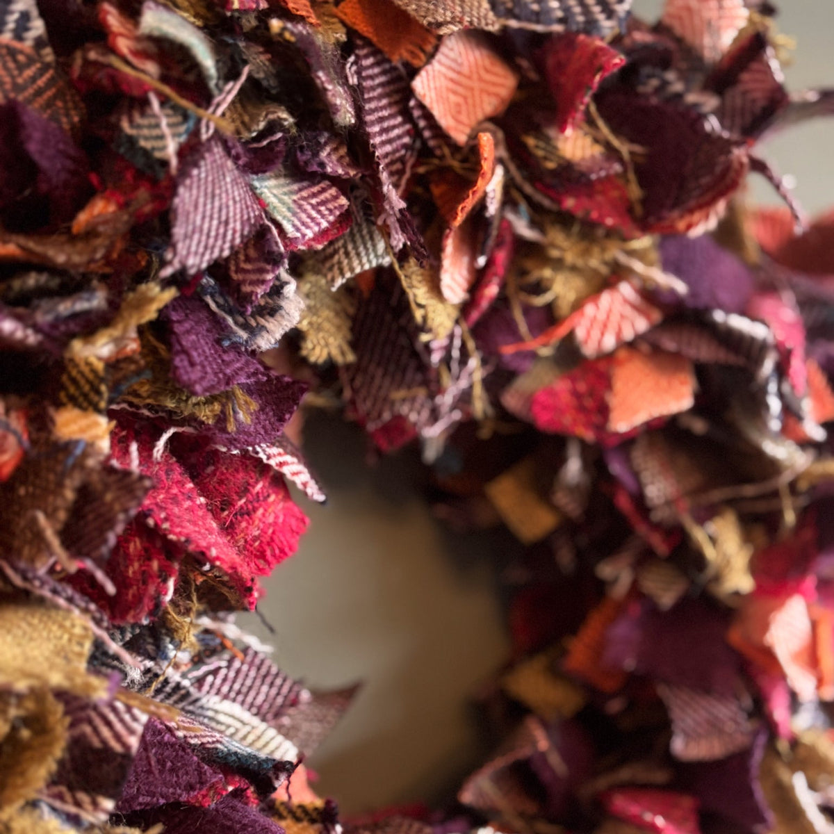 A close-up of a handmade fabric wreath made from layered, frayed plaid and tweed fabric strips in autumnal colors including burgundy, rust orange, mustard yellow, and brown.