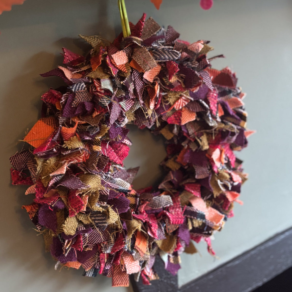 A handmade autumn-themed fabric wreath hanging on a gray wall below a wooden mantel. The wreath is made of layered, fringed fabric strips in shades of burgundy, rust orange, terracotta, and brown.