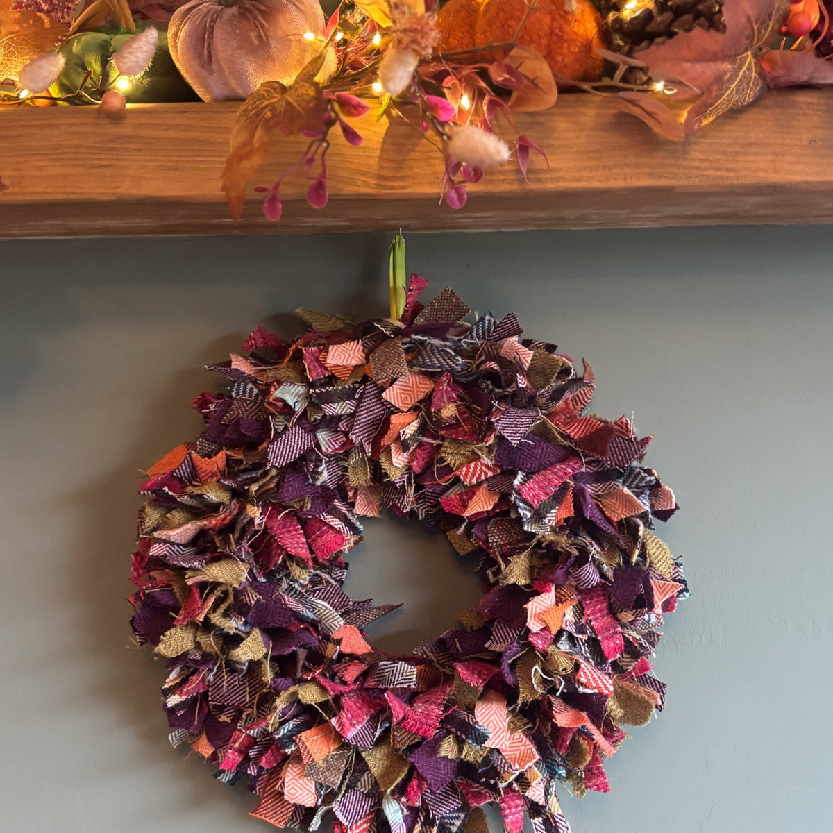 A handmade autumn-themed fabric wreath hanging on a dark wall below a wooden mantel decorated with pumpkins, pinecones, and fall leaves with warm string lights.