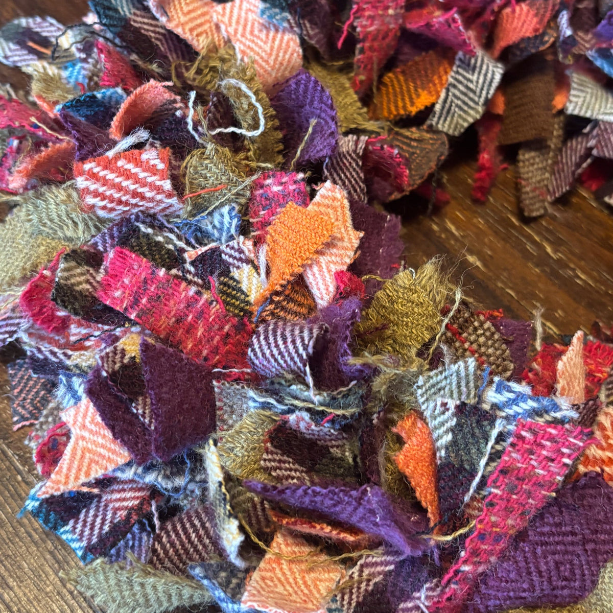A handmade fabric wreath made from multi-colored plaid and tweed fabric strips, resting on a rustic wooden surface.