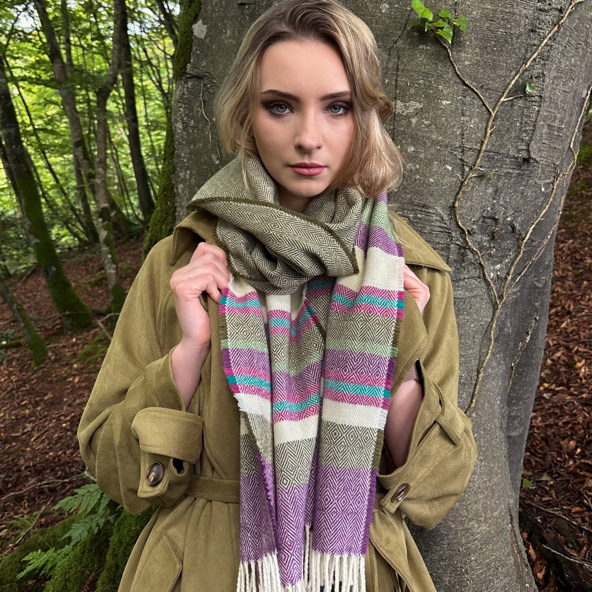 A woman wearing a thick striped knit scarf in olive green, purple, pink, and cream, styled with an olive green coat and standing in front of a large tree trunk in a forest.