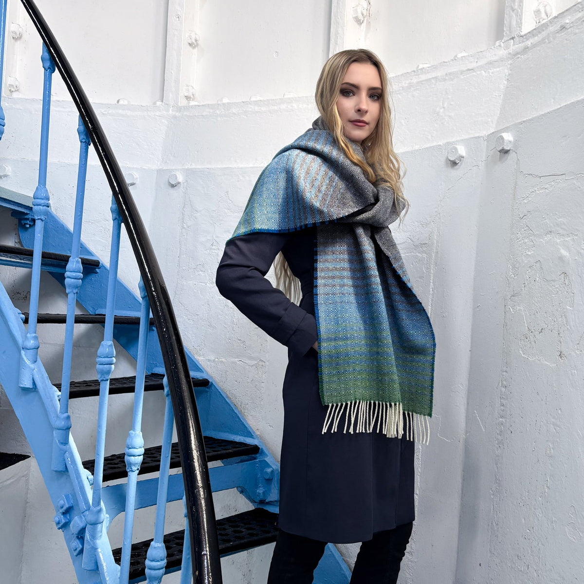 A woman wearing a long navy coat and black pants stands on a blue spiral staircase, wrapped in a large patterned scarf with blue, green, and cream tones and fringe ends.