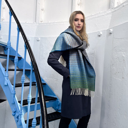 A woman wearing a long navy coat and black pants stands on a blue spiral staircase, wrapped in a large patterned scarf with blue, green, and cream tones and fringe ends.