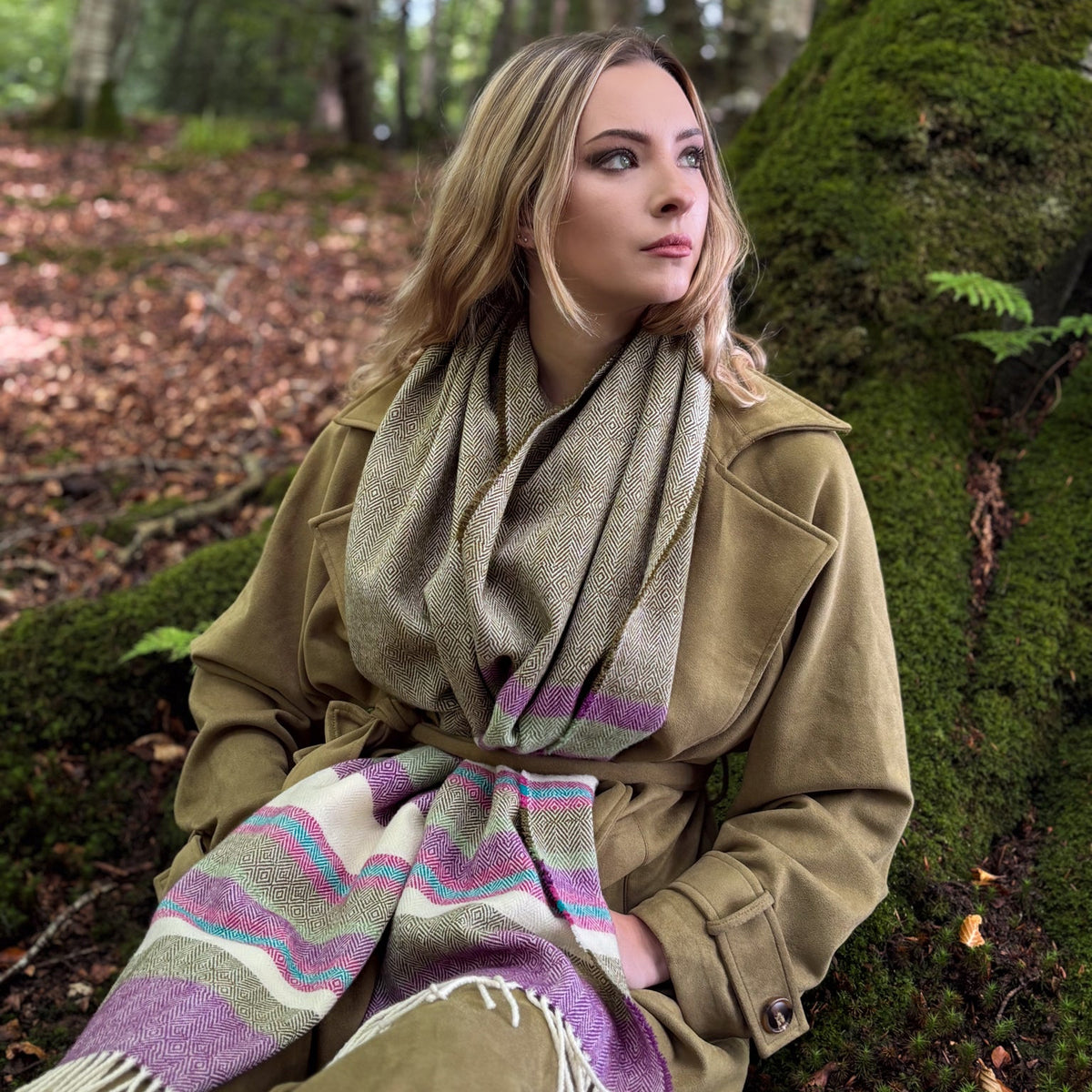 A woman sitting in a mossy forest wearing a tan coat and a striped wool scarf in beige, purple, pink, and green.