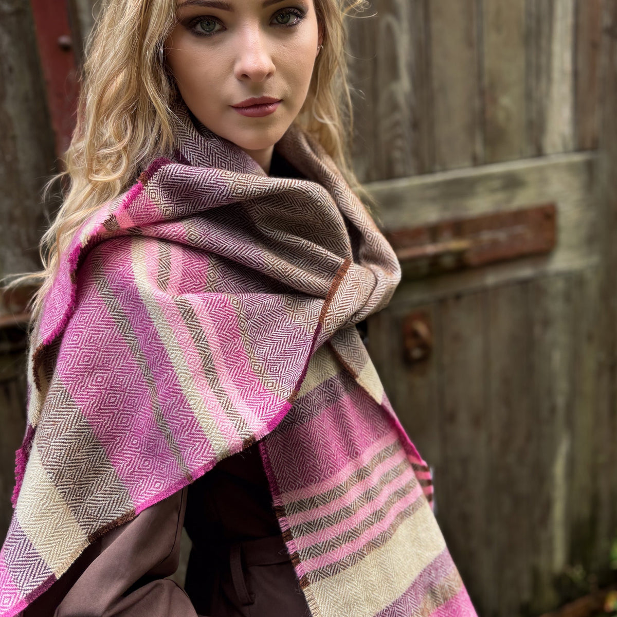 A woman wearing a pink, brown, and beige patterned woven shawl or wrap draped over her shoulders, posed in front of a rustic wooden door.