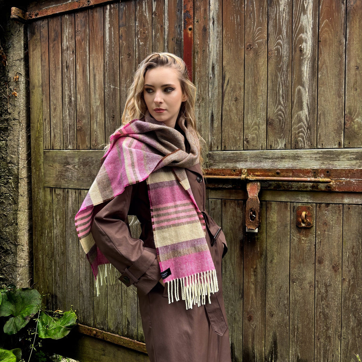 A woman stands in front of weathered wooden barn doors wearing a brown coat and a pink, beige, and gray plaid fringed scarf.