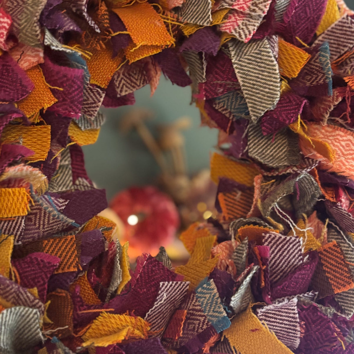 A close-up of a handmade fabric wreath made from layered, frayed woven strips in autumn colors including burgundy, orange, mustard yellow, brown, and gray, with a soft blurred background.
