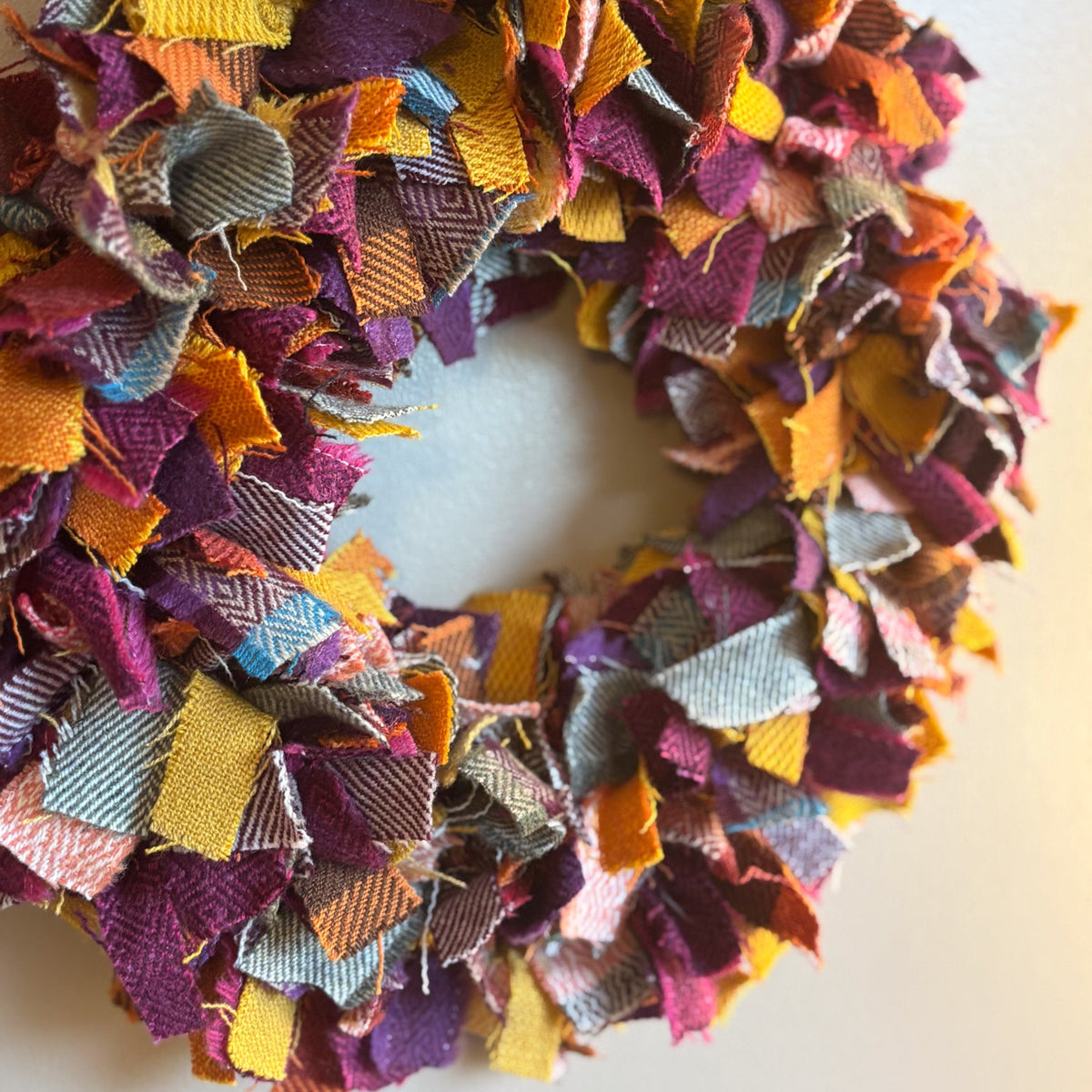 A handmade fabric wreath made from layered, frayed textile pieces in autumn colors including purple, orange, yellow, pink, and gray, hanging against a plain off-white wall.