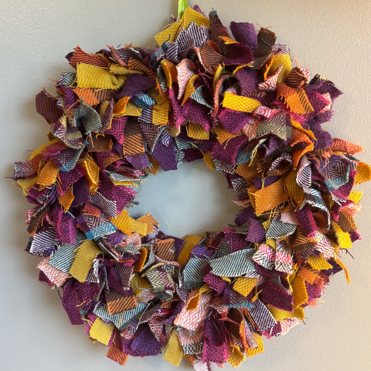 A handmade fabric fringe wreath hanging on a plain light gray wall. The wreath is made of many overlapping ribbon-like strips in assorted colors including purple, orange, yellow, pink, blue, and gray, with a green hanging loop at the top.