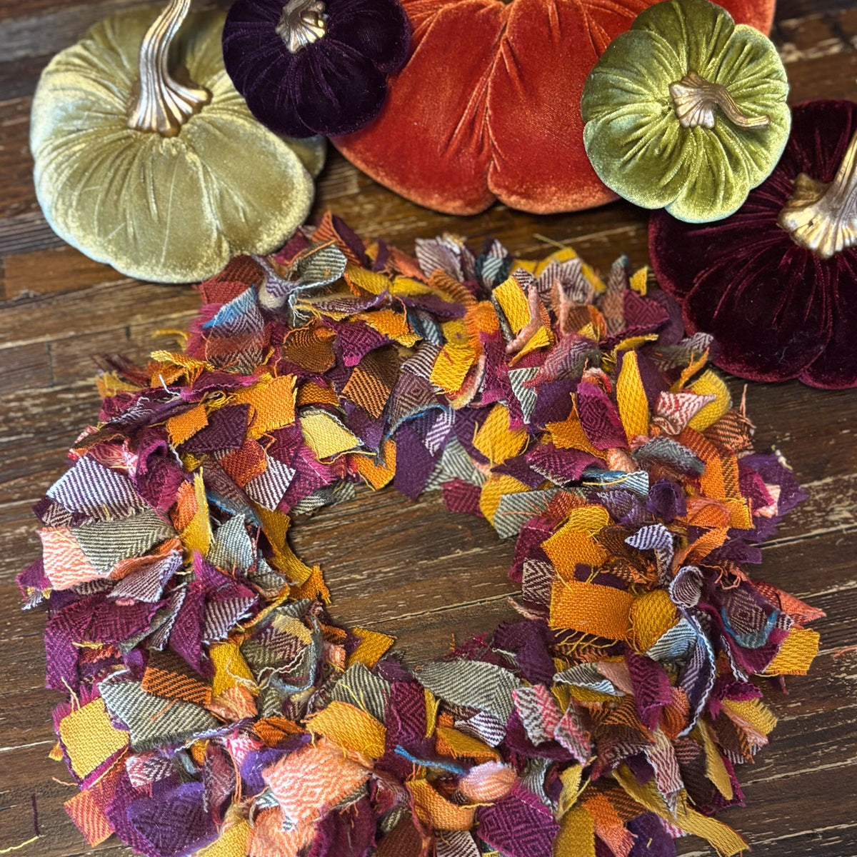 A handmade autumn-themed fabric wreath made of colorful frayed ribbon in shades of orange, purple, yellow, and pink, displayed on a dark wooden surface with velvet pumpkins in the background.