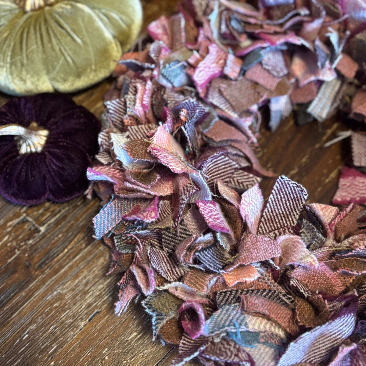 A close-up of a fabric flower wreath made from layered, ruffled petals in muted pink, mauve, brown, and gray tones, arranged on a rustic wooden surface with velvet pumpkins in the background.