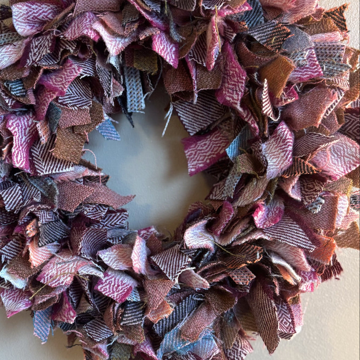A handmade fabric wreath made from layered, frayed ribbon strips in dusty pink, mauve, purple, brown, and muted blue, hanging against a plain light gray wall.