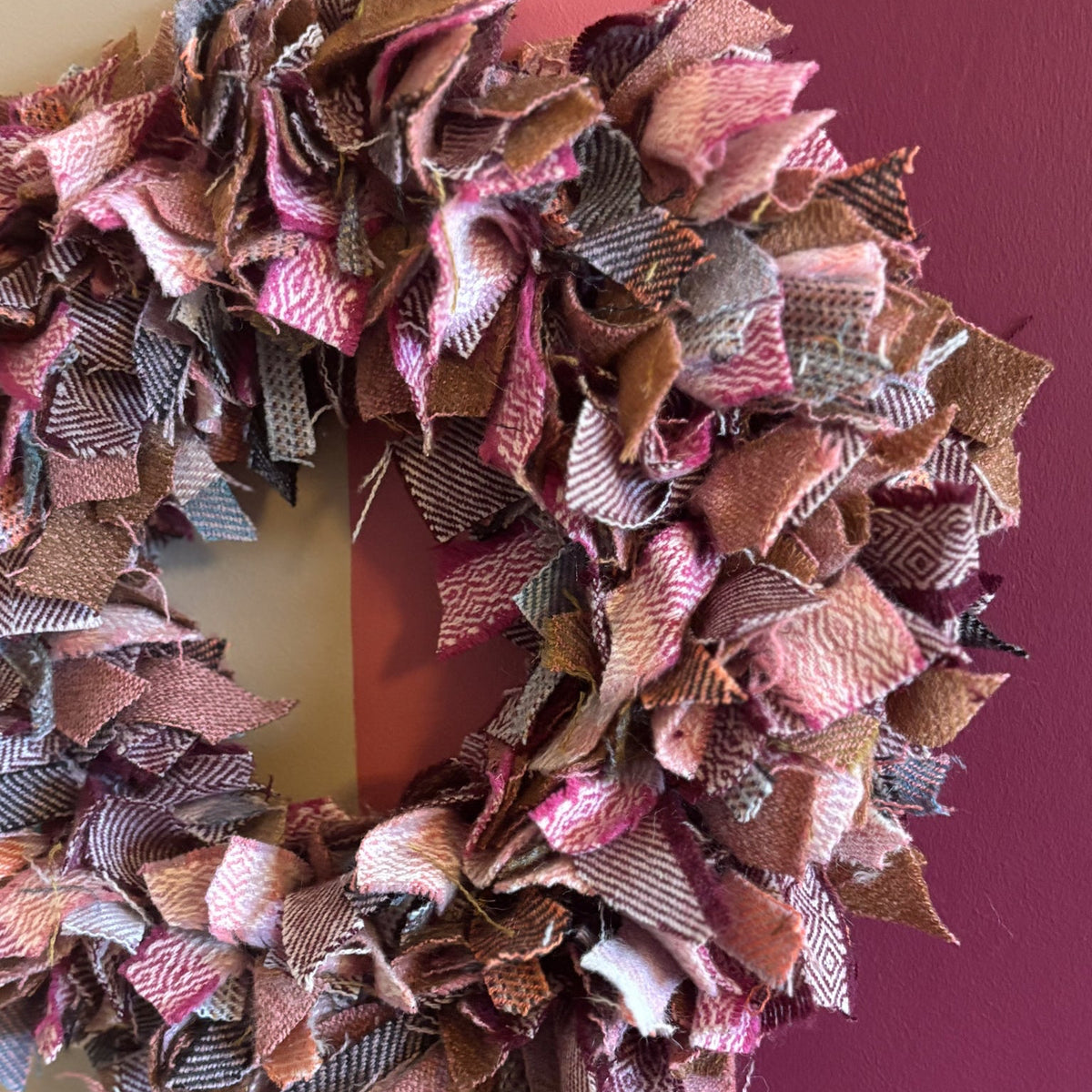 A handmade fabric slub wreath made from layered ribbon-like strips in pink, brown, beige, and gray tones, hanging against a painted wall with cream, pink, and dark purple sections.