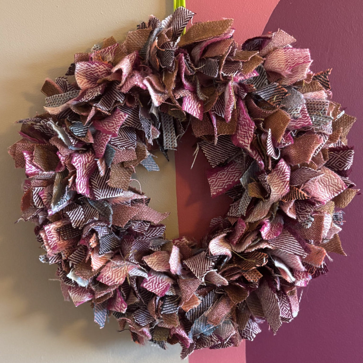 A handmade fabric ribbon wreath hanging on a wall, featuring layered ruffles in dusty pink, mauve, brown, and gray tones, with a bright green hanging loop at the top.