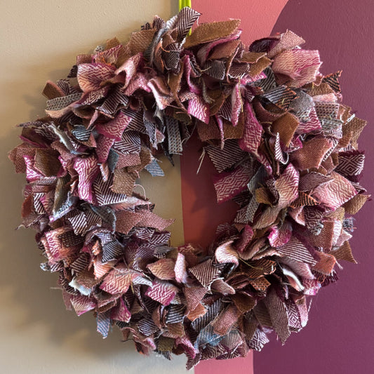 A handmade fabric ribbon wreath hanging on a wall, featuring layered ruffles in dusty pink, mauve, brown, and gray tones, with a bright green hanging loop at the top.
