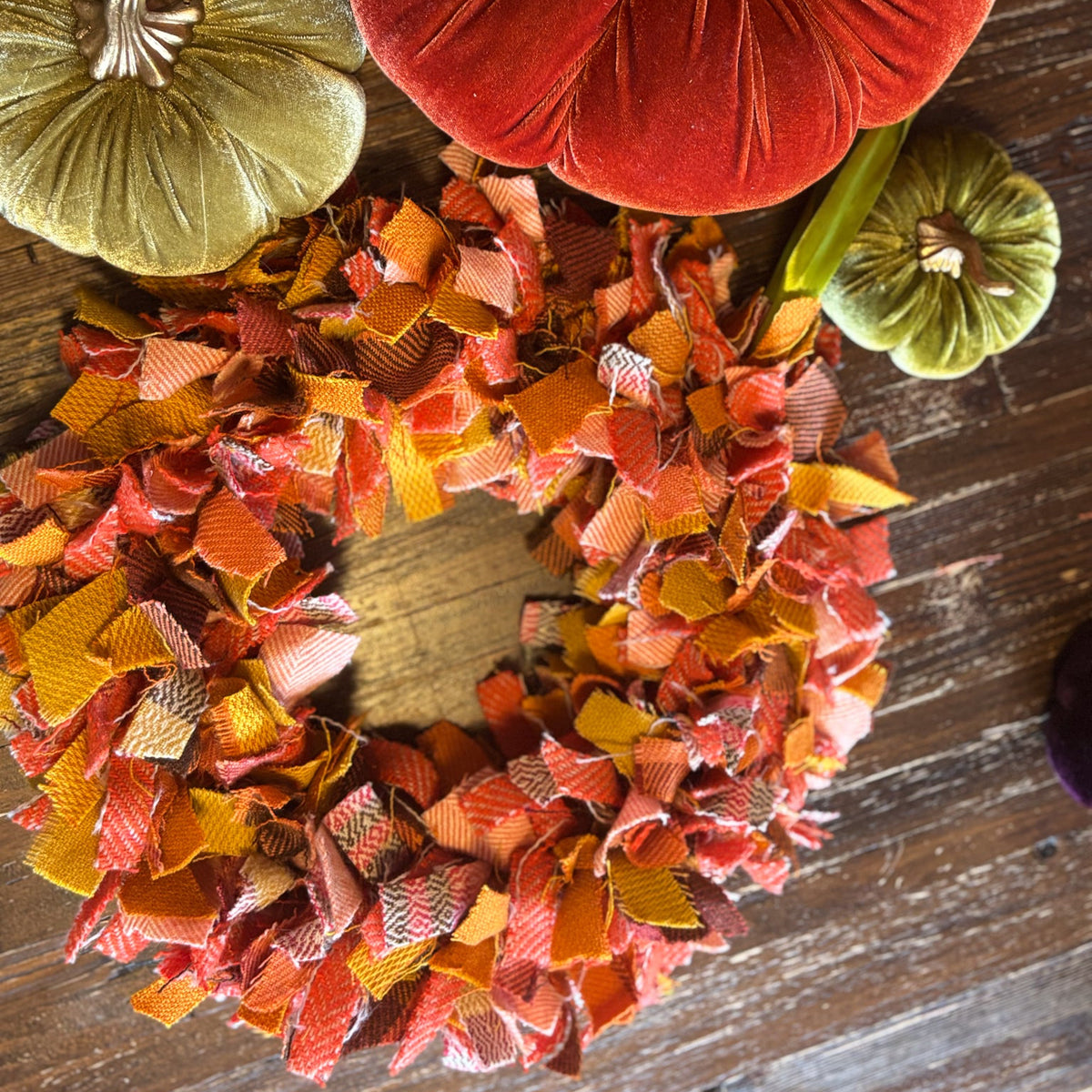An autumn-themed fabric wreath made of layered ribbon strips in orange, red, yellow, and brown tones, displayed on a rustic wooden surface alongside decorative velvet pumpkins.