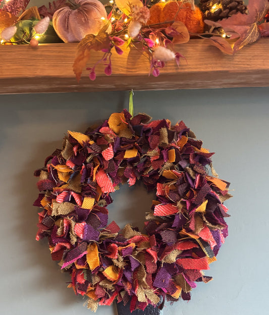 A handmade autumn-themed fabric wreath hanging on a light blue-gray wall below a wooden mantel decorated with fall leaves, pumpkins, and string lights.