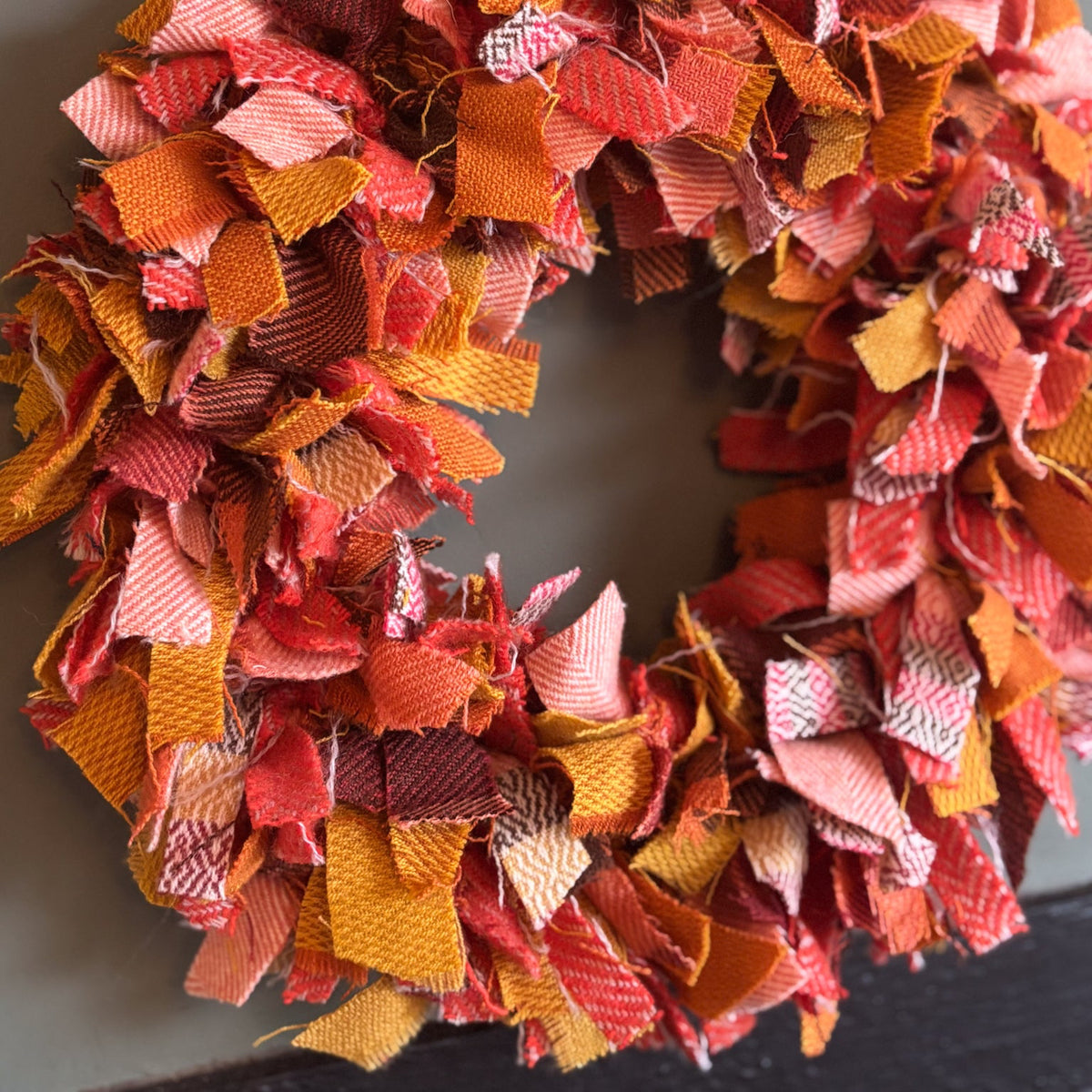 A handmade autumn-themed fabric shag wreath made from colorful patterned fabric scraps in shades of orange, red, pink, and yellow, hanging against a dark gray wall.