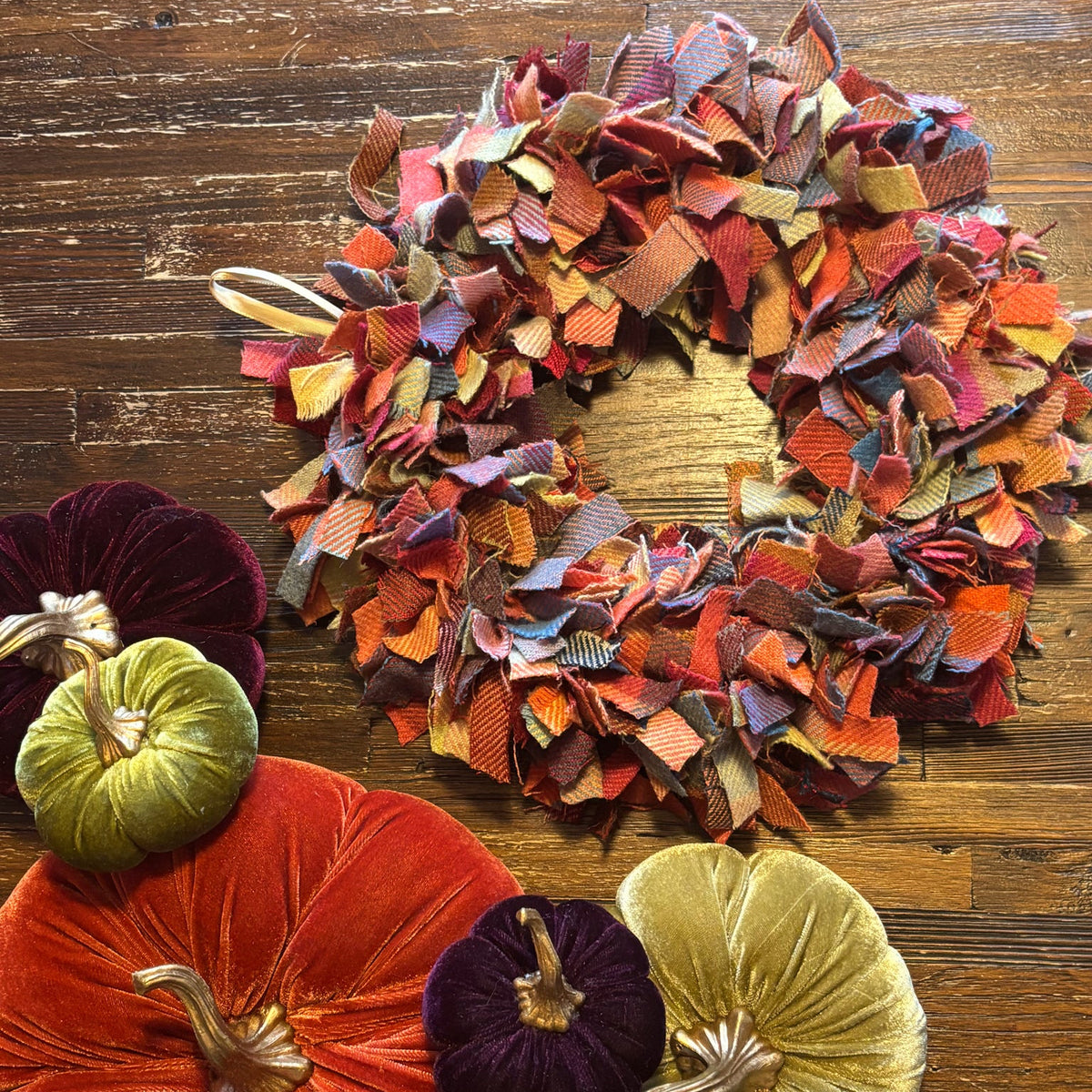 A rustic autumn-themed decorative wreath made of layered ribbon strips in fall colors, displayed on a dark wood surface alongside velvet pumpkins in orange, gold, purple, and green.