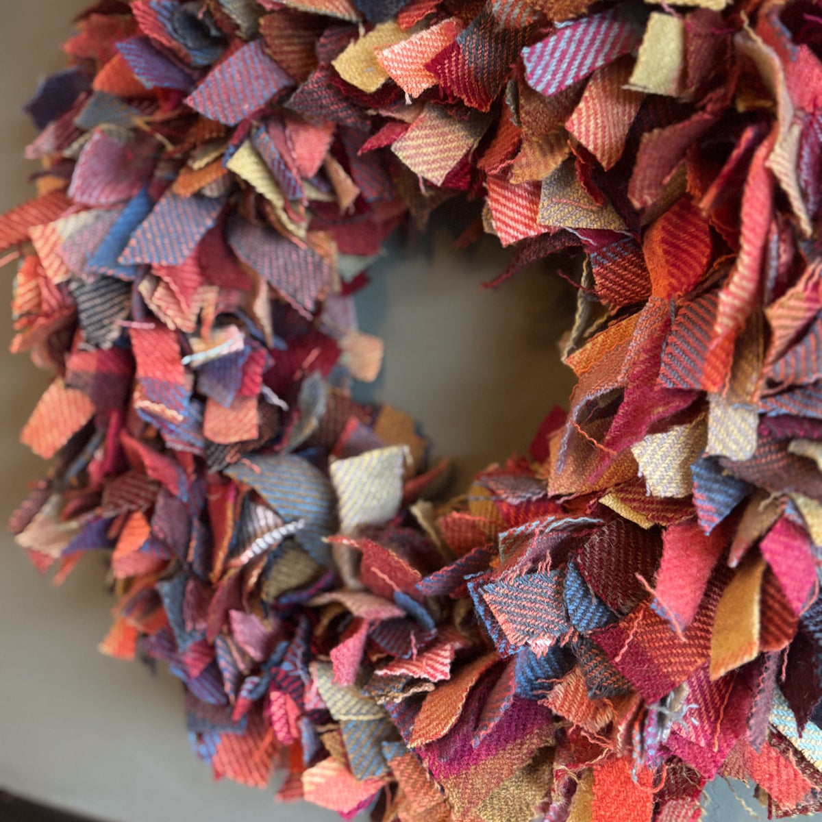 A handmade fabric wreath made from many small plaid and striped cloth strips in autumnal colors including red, orange, blue, purple, pink, and beige, photographed close up against a neutral gray background.