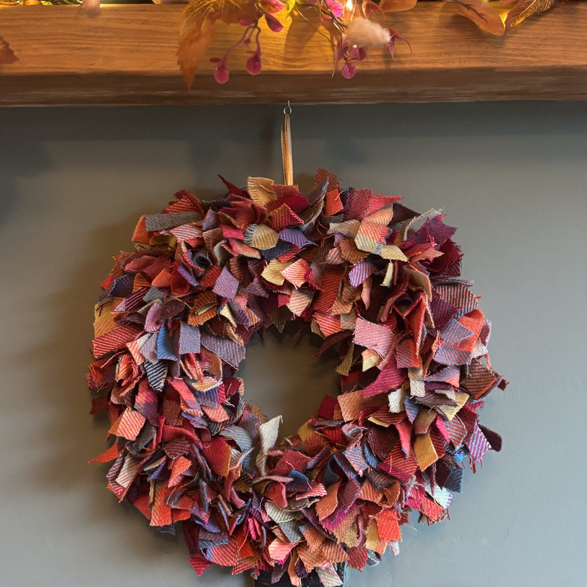 A handmade autumn-themed fabric wreath made from layered, fringed fabric strips in shades of red, pink, orange, purple, and blue, hanging on a gray wall below a wooden mantel decorated with pumpkins, pinecones, and fall leaves with warm string lights.