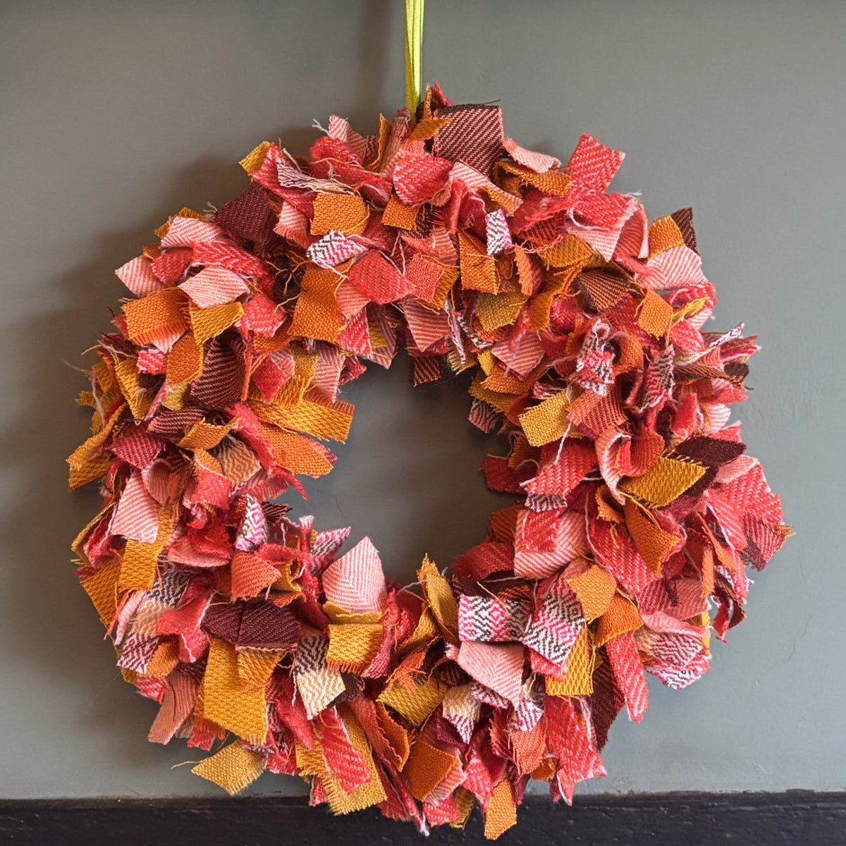 An autumn-themed fabric fringe wreath in shades of orange, red, pink, and yellow hangs on a dark gray wall below a wooden mantel decorated with fall leaves, pumpkins, and string lights.