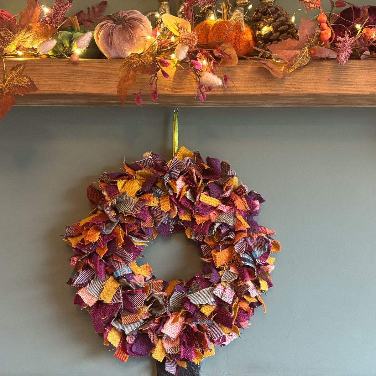An autumn-themed fabric wreath in orange, purple, and yellow hangs on a light blue-gray wall below a wooden mantel decorated with fall leaves, pumpkins, and warm string lights.