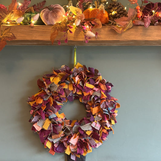 An autumn-themed fabric wreath in orange, purple, and yellow hangs on a light blue-gray wall below a wooden mantel decorated with fall leaves, pumpkins, and warm string lights.