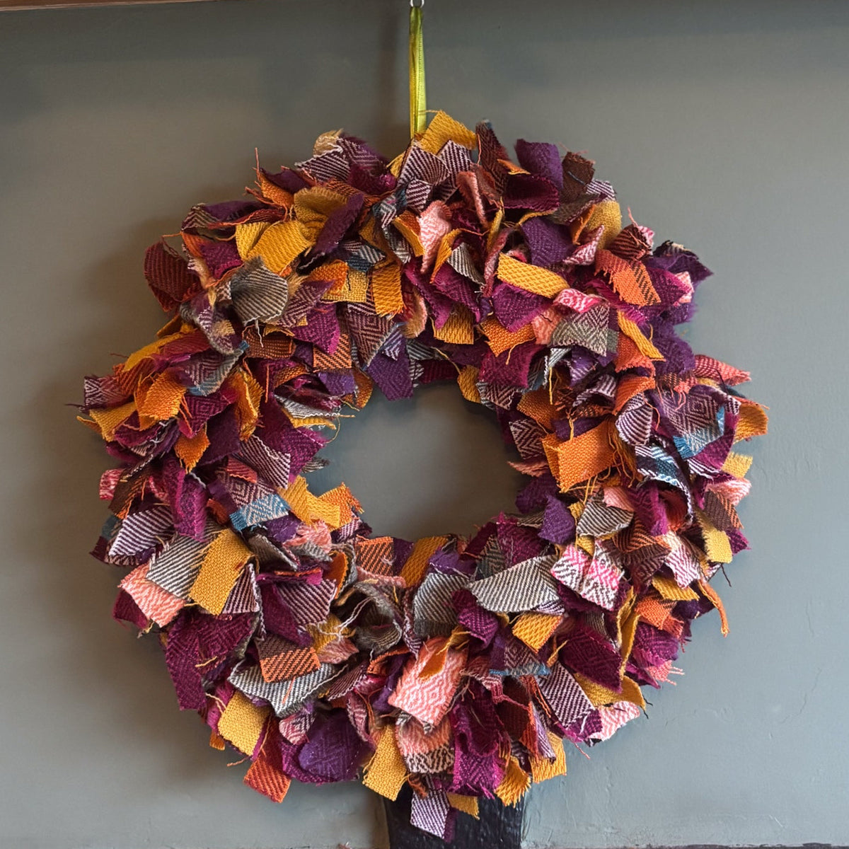 A handmade autumn-themed fabric wreath made of layered ruffled strips in orange, purple, pink, and brown, hanging on a gray wall below a decorated wooden mantel with pumpkins and fairy lights.