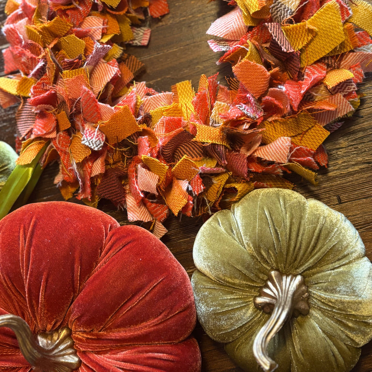 A handmade autumn-themed fabric wreath made from colorful plaid and patterned fabric strips in orange, red, yellow, and brown, displayed on a dark wooden surface next to two velvet pumpkins.