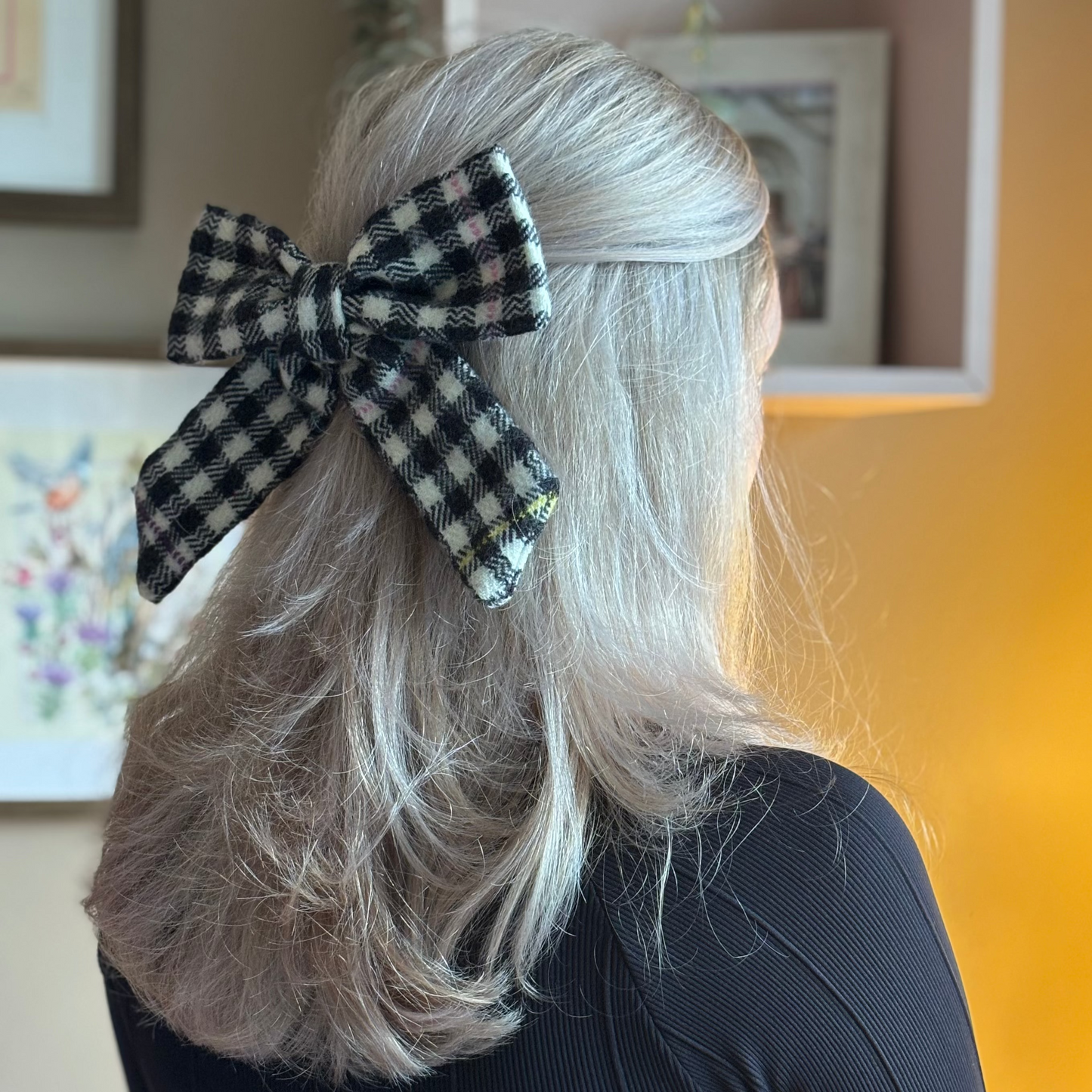 Person with a checkered Tweed bow in their hair, wearing a black top standing in a room with framed pictures on the wall.