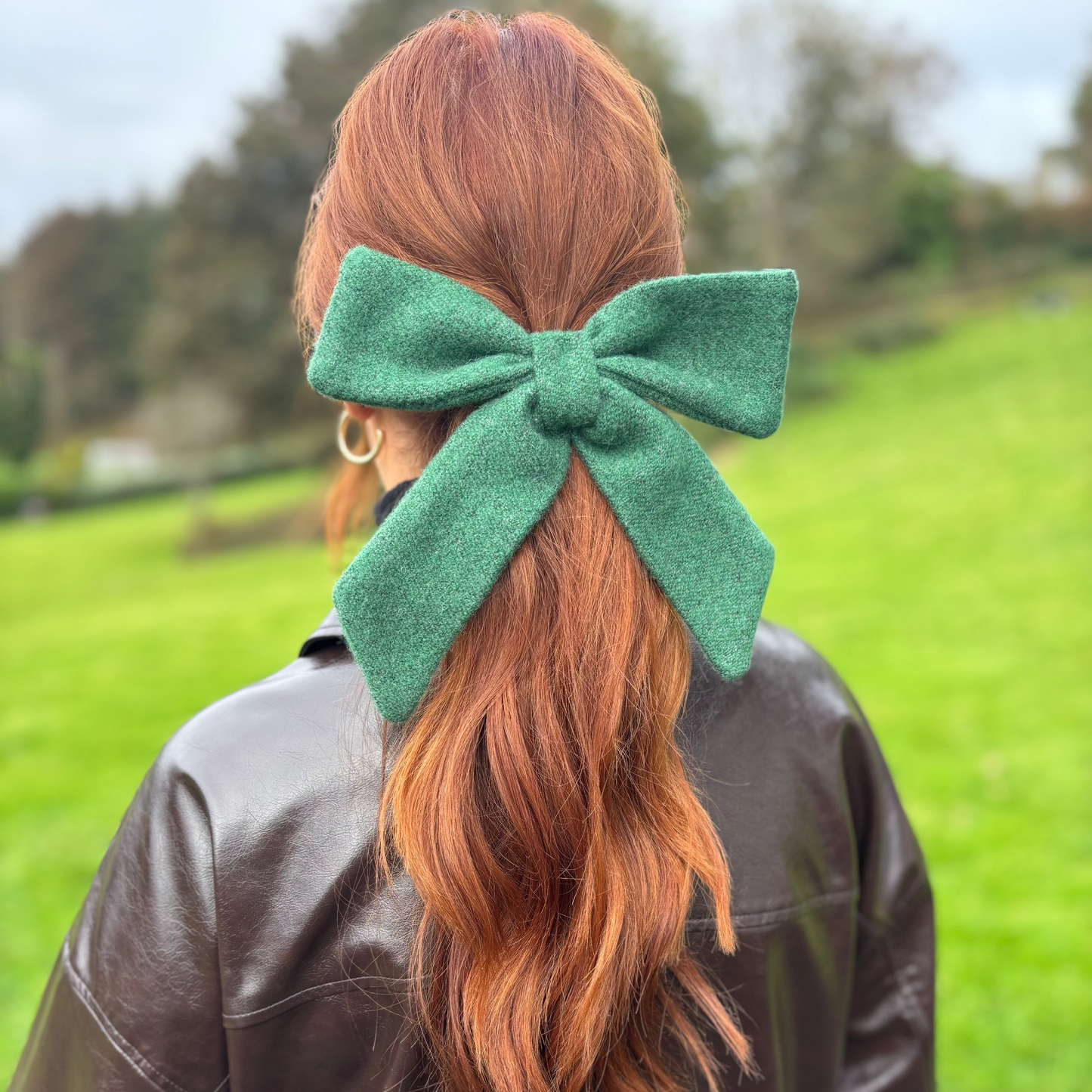 Person with red hair tied in a ponytail with a green Tweed bow, wearing a brown leather jacket outdoors.