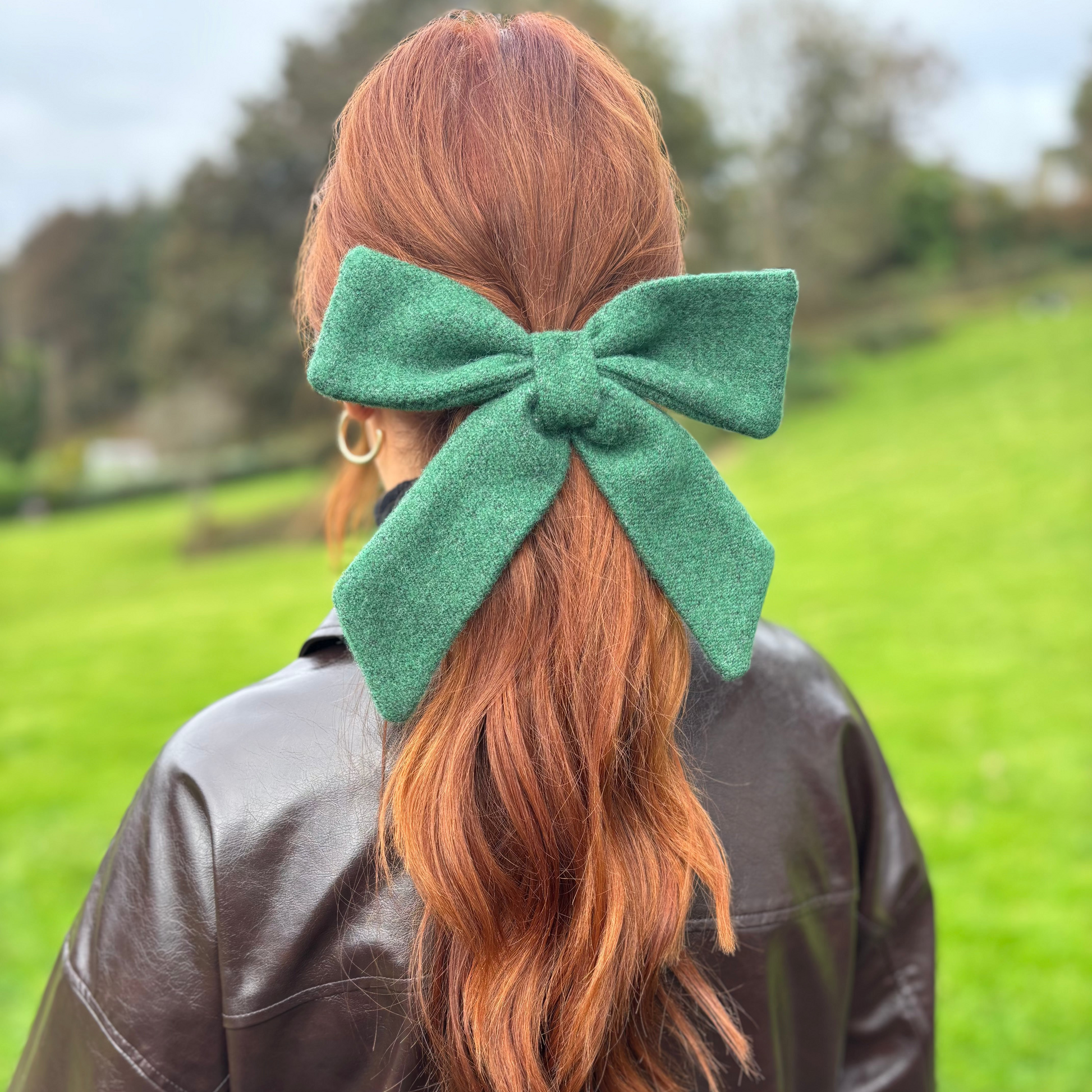 Person with red hair tied in a ponytail with a green Tweed bow, wearing a brown leather jacket outdoors.