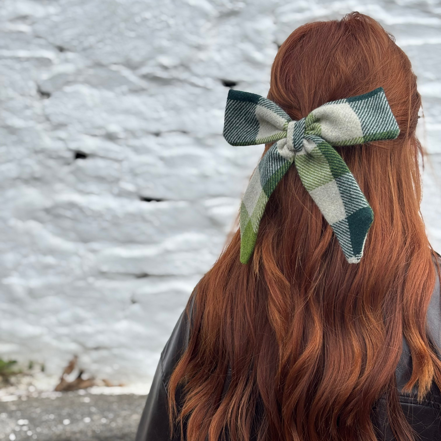 Person with long red hair wearing a green plaid/Tweed bow tie against a textured wall.