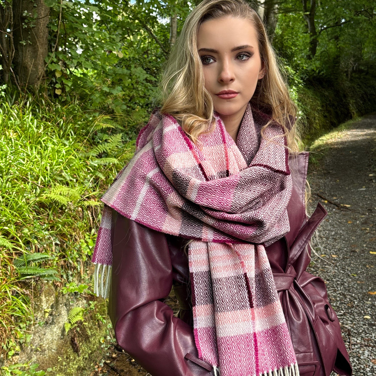 A woman standing on a forest path wearing a burgundy faux-leather coat and a pink and white plaid scarf.