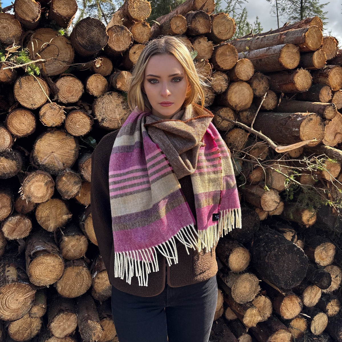 A woman stands outdoors in front of a large stack of cut logs, wearing a multicolor plaid scarf with white fringe. The scarf features stripes of pink, purple, brown, beige, and gray.