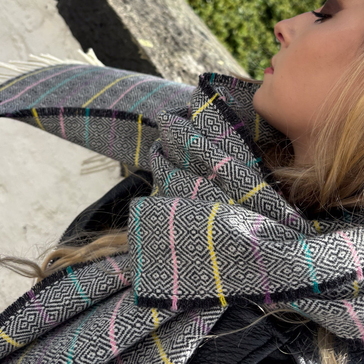 A close-up lifestyle shot of a woman wearing a black and gray geometric patterned scarf with thin multicolor stripes of pink, yellow, and blue.