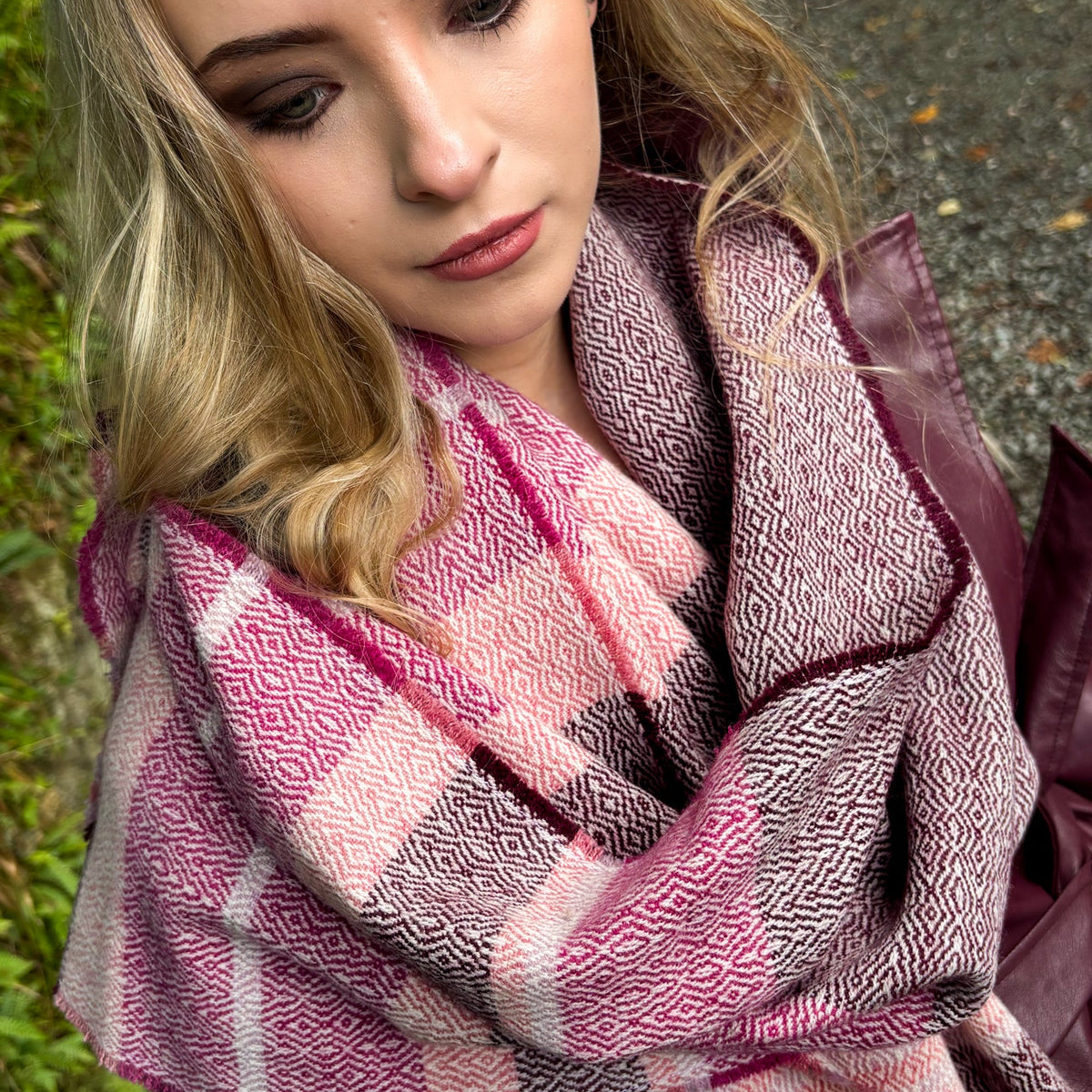 A close-up lifestyle shot of a woman wearing a pink, purple, and white patterned scarf wrapped around her neck, with a dark burgundy jacket visible on the side.
