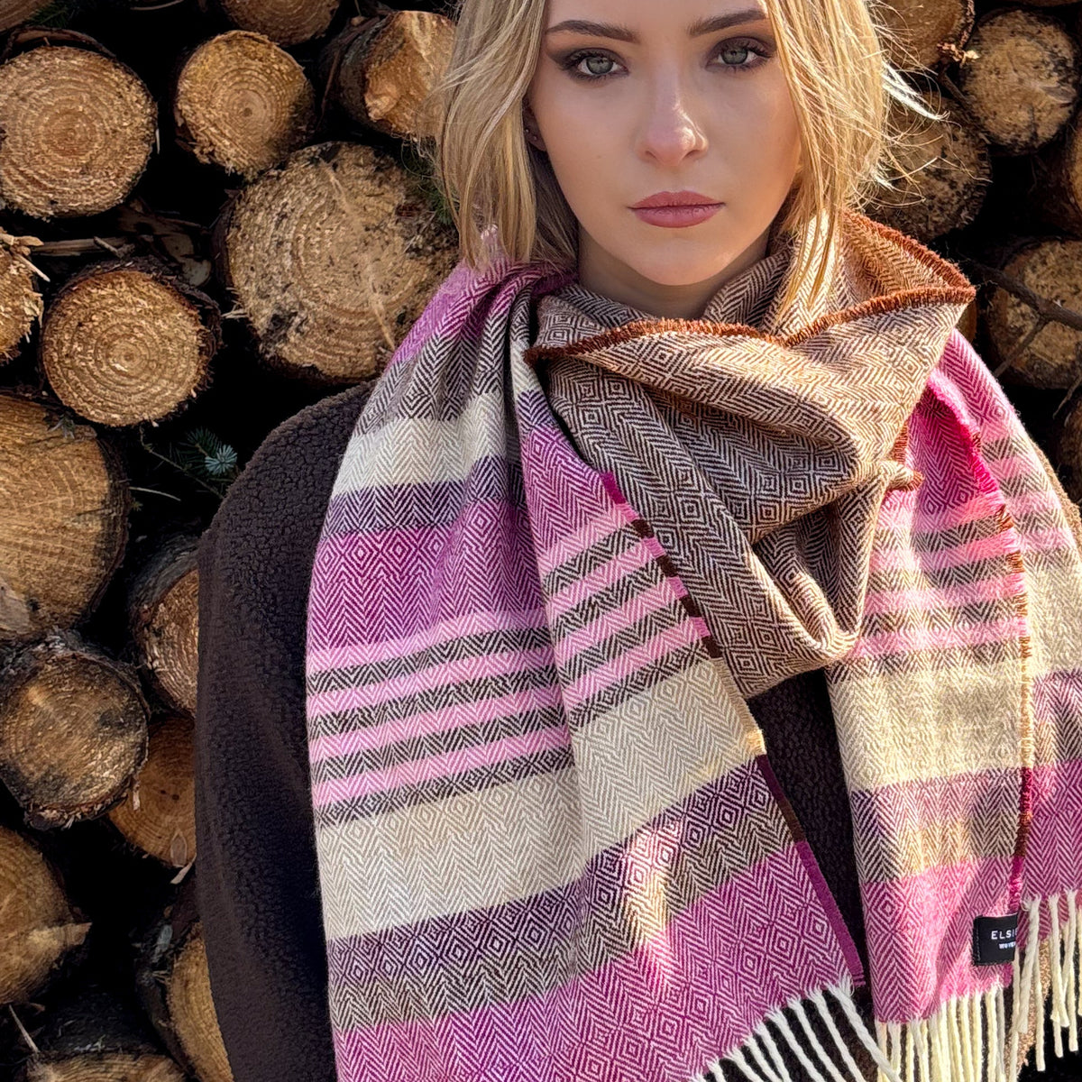 A woman wearing a thick woven scarf with pink, beige, and brown herringbone and striped patterns, posed in front of a stack of cut logs.