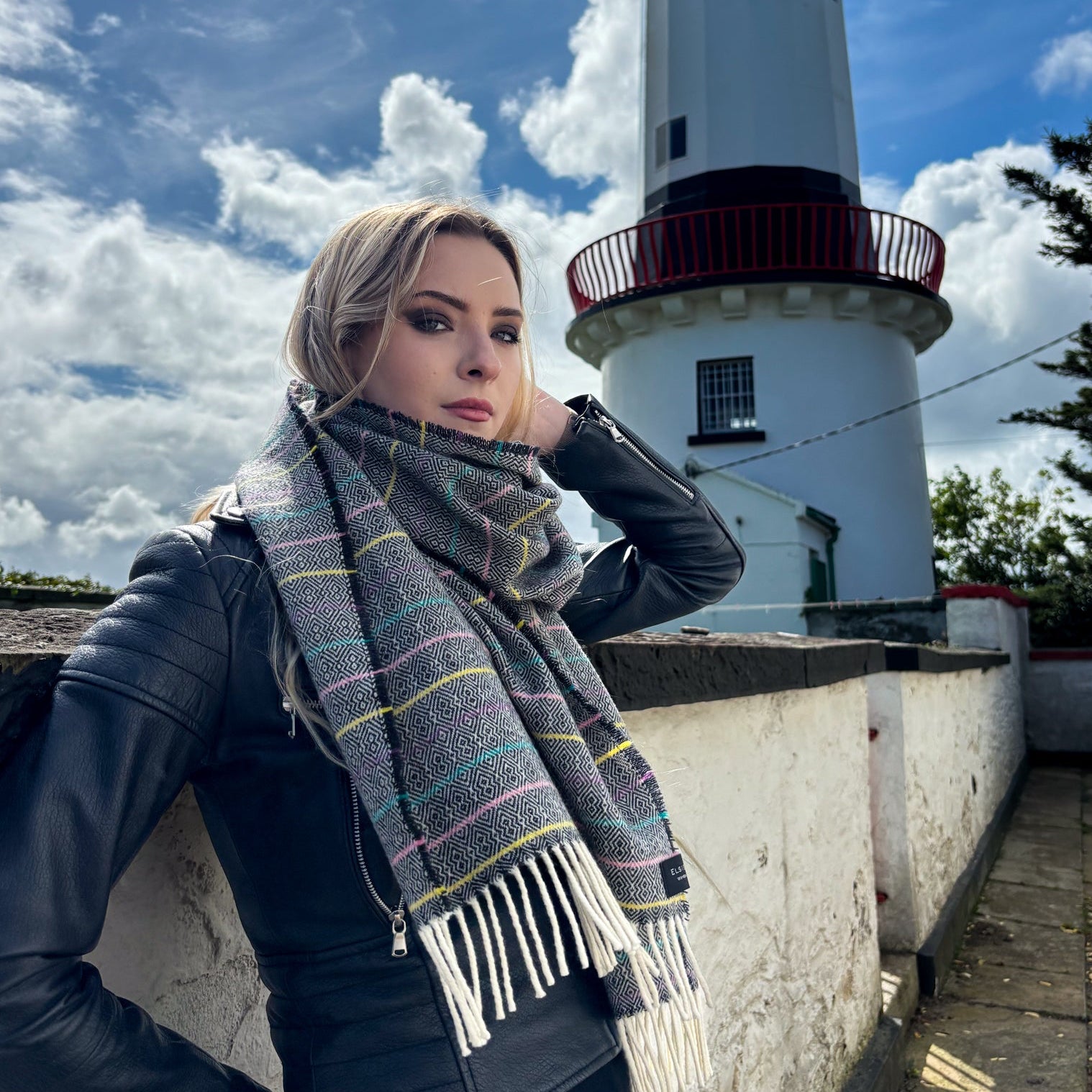 A woman wearing a gray striped knit scarf with white fringe and a black leather jacket stands outdoors in front of a white lighthouse under a blue sky with clouds.