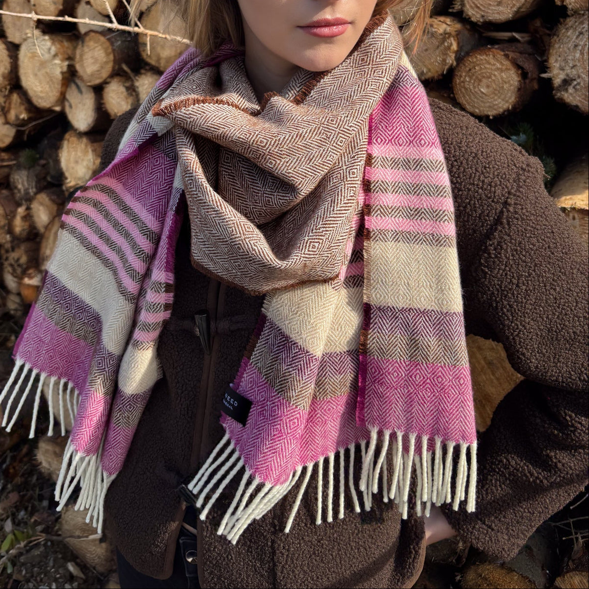A woman wearing a thick woven scarf with pink, beige, and brown stripes and white fringe, standing in front of a stacked woodpile outdoors.