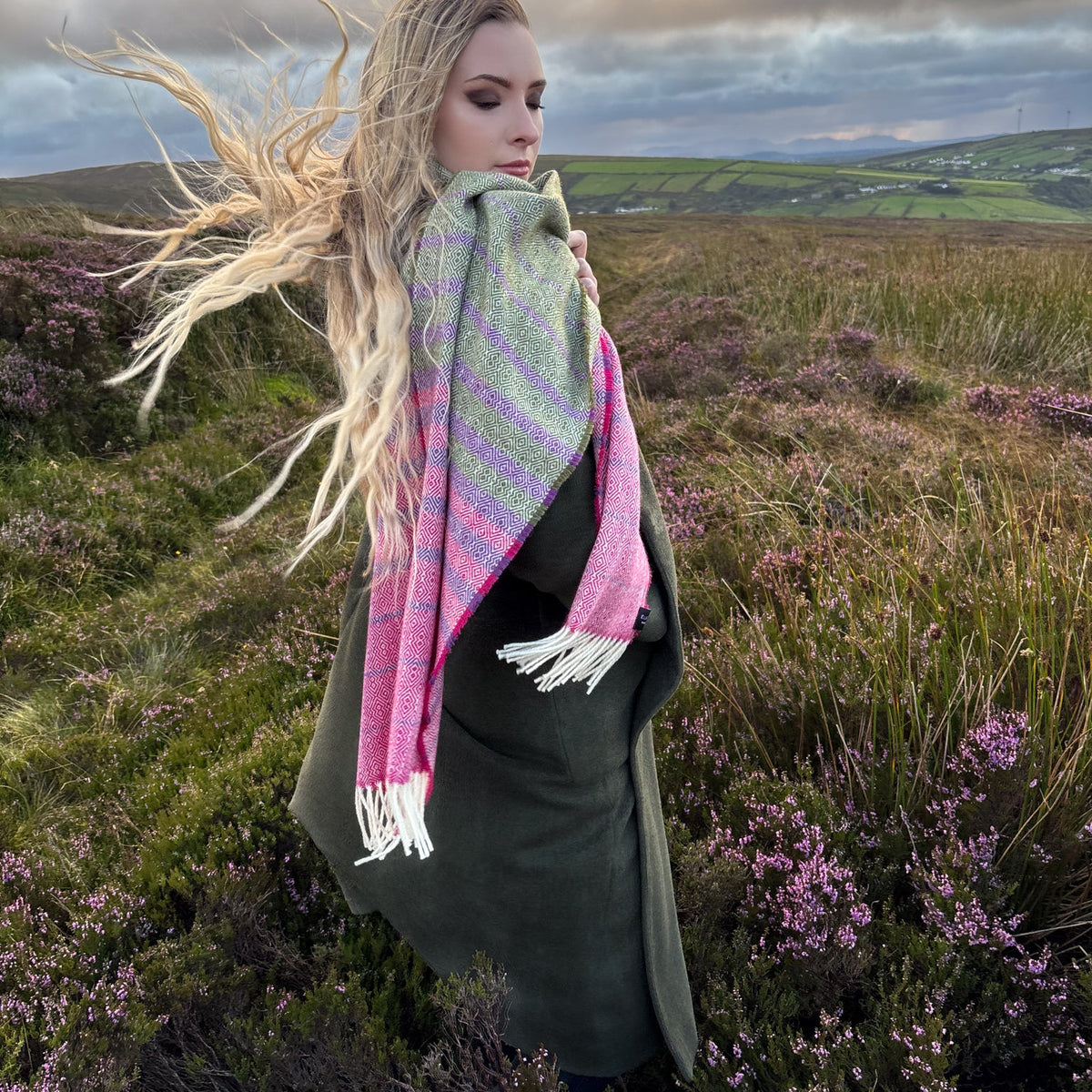 A woman standing in an outdoor heather hill in Ireland  under a dramatic cloudy sky, wearing a olive green pink and purple wrap with white fringe.