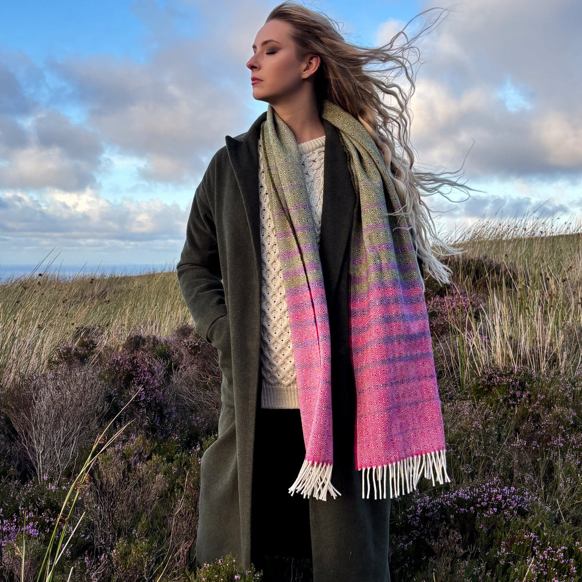 A woman standing outdoors in a grassy field under a cloudy blue sky, wearing a long olive green coat and woven scarf with a green, pink, and purple striped pattern and white fringe.