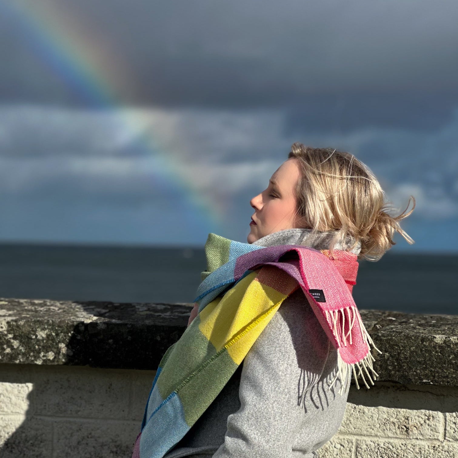 Person wearing a colorful rainbow tweed scarf with a irish rainbow in the background