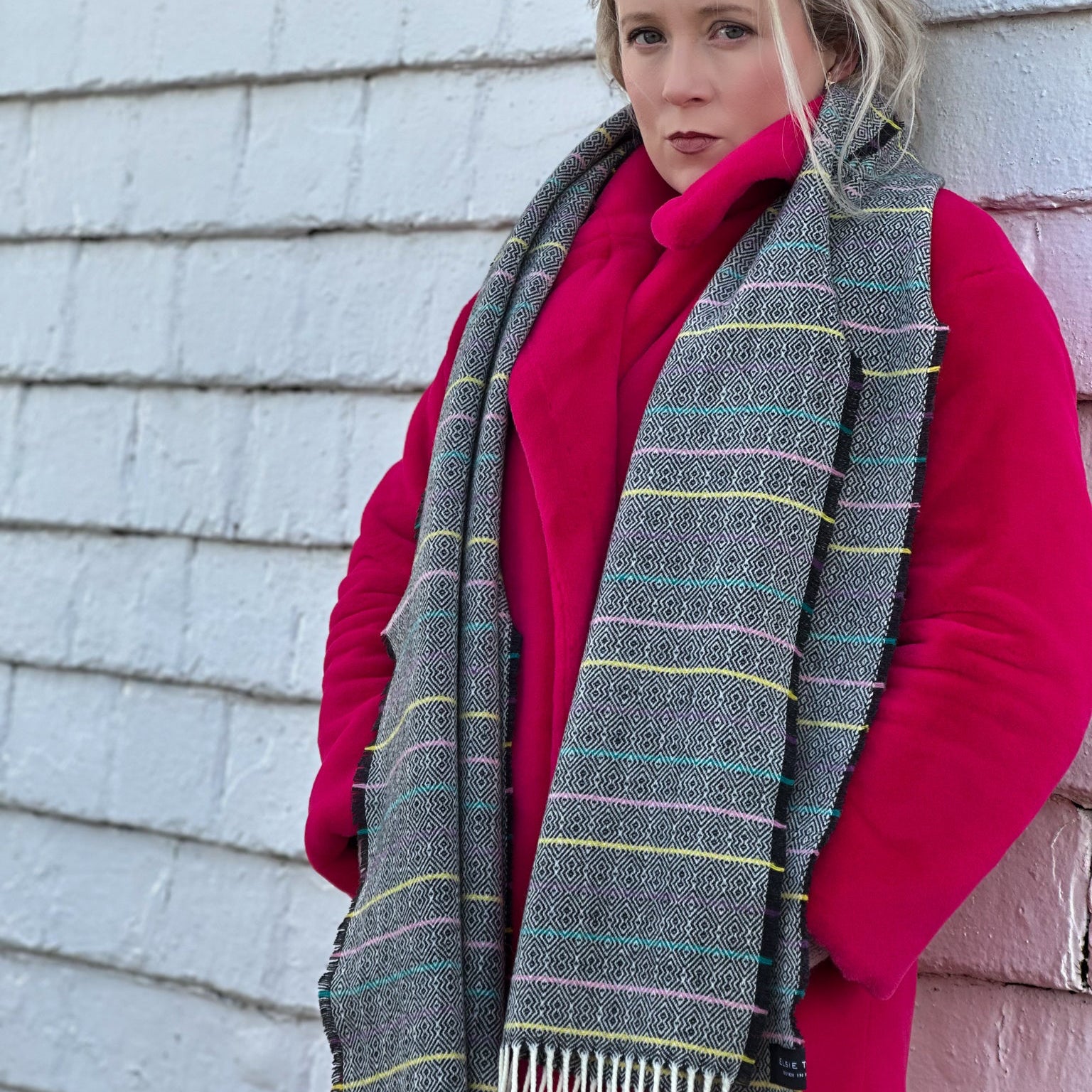 Woman wearing a pink coat and black striped scarf against a light-colored wall.