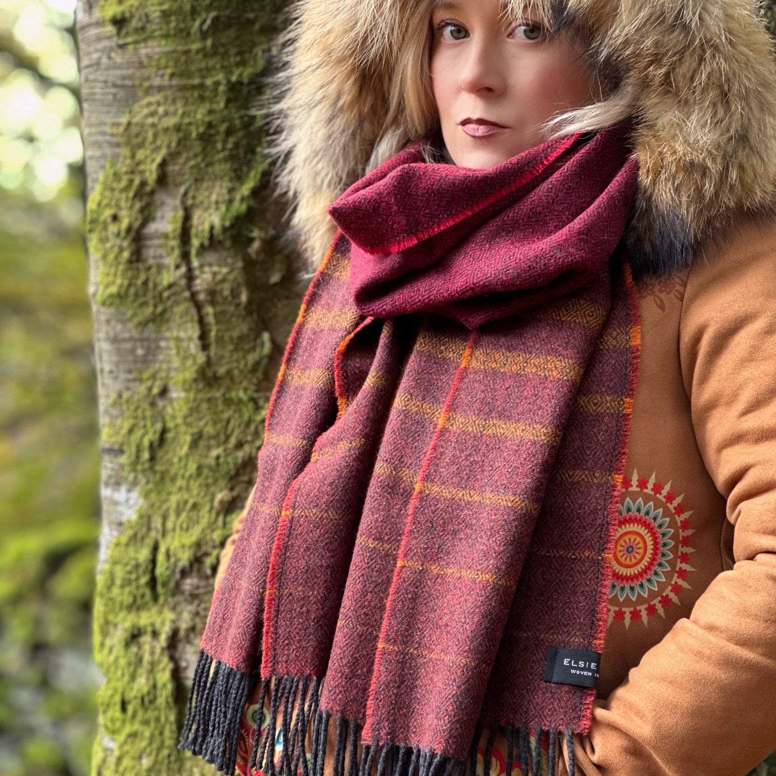 Person wearing a plaid scarf and brown coat with fur hood in a forest setting
