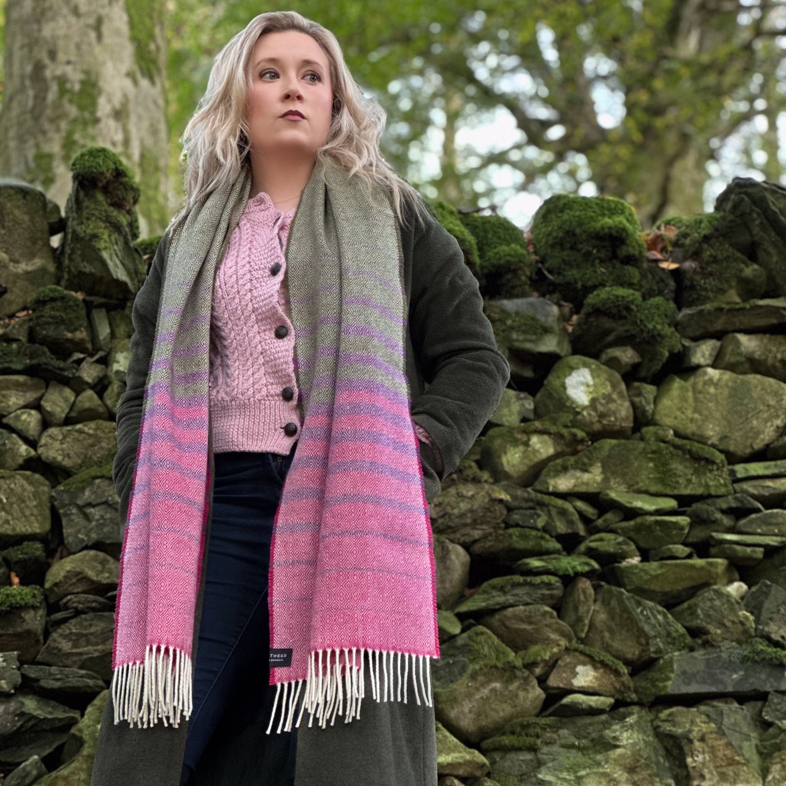Woman wearing a pink and gray striped scarf standing in front of a stone wall with trees in the background